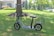 An electric scooter with a black frame and a seat is parked on a grassy lawn. It features a wire basket attached to the rear. The background includes a tree, surrounding greenery, and a residential house partially visible with a cream facade and a window.