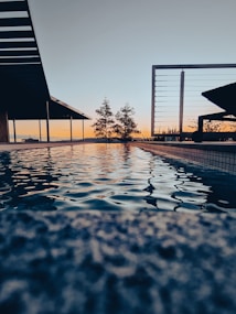 A serene outdoor pool reflecting the colors of a sunset sky, surrounded by modern architectural elements and minimal landscaping. The surface of the water ripples gently, capturing the warm hues of orange and blue from the horizon.