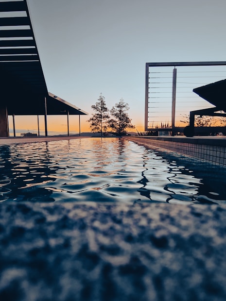 A serene outdoor pool reflecting the colors of a sunset sky, surrounded by modern architectural elements and minimal landscaping. The surface of the water ripples gently, capturing the warm hues of orange and blue from the horizon.