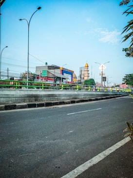 A cityscape featuring a busy urban road lined with greenery and streetlights. In the background, various commercial buildings display advertisements, and a mosque with a golden dome rises prominently. The sky is clear and blue, adding a vibrant contrast to the scene.
