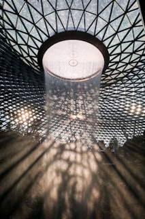 A large indoor waterfall cascades from the center of a geometric glass dome, creating dramatic light patterns and shadows on the surrounding area. The architectural structure is intricate, featuring a lattice design with triangular shapes that allow diffused sunlight to shine through.