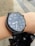 A black smartwatch is displayed on a person's wrist, covered in raindrops. The screen shows the time as 08:33 with additional information like the date and a step counter. The watch has a glossy, circular display and appears to be in an outdoor setting with a blurred background.