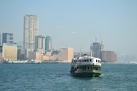 A ferry is traveling across a body of water with a backdrop of a cityscape featuring modern skyscrapers and construction cranes. The city appears bustling, with a mix of tall buildings, some of which have recognizable brand names.