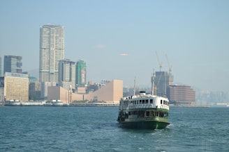 A ferry is traveling across a body of water with a backdrop of a cityscape featuring modern skyscrapers and construction cranes. The city appears bustling, with a mix of tall buildings, some of which have recognizable brand names.