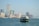 A ferry is traveling across a body of water with a backdrop of a cityscape featuring modern skyscrapers and construction cranes. The city appears bustling, with a mix of tall buildings, some of which have recognizable brand names.