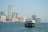 A ferry is traveling across a body of water with a backdrop of a cityscape featuring modern skyscrapers and construction cranes. The city appears bustling, with a mix of tall buildings, some of which have recognizable brand names.
