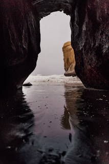 A view through a dark cave opening reveals the ocean beyond, with a notable rock formation in the distance partially covered by mist. The cave floor is wet, reflecting the light from outside, adding a sense of mystery and depth to the scene.
