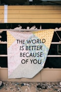 A patchwork quilt with a variety of floral patterns is draped over a broken window of an abandoned building. The quilt prominently displays the message 'THE WORLD IS BETTER BECAUSE OF YOU' in bold black letters. Broken glass is scattered on the ground in front of the window, adding contrast to the colorful quilt.