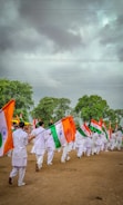 A group of people dressed in white uniforms are marching on a dirt ground holding Indian flags. The background features lush green trees under a cloudy sky.