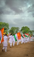 A group of people dressed in white uniforms are marching on a dirt ground holding Indian flags. The background features lush green trees under a cloudy sky.