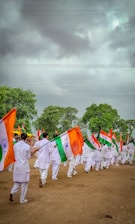 A group of people dressed in white uniforms are marching on a dirt ground holding Indian flags. The background features lush green trees under a cloudy sky.