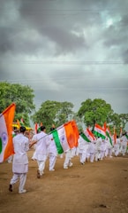 A group of people dressed in white uniforms are marching on a dirt ground holding Indian flags. The background features lush green trees under a cloudy sky.