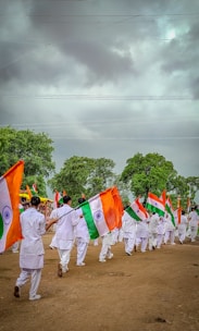 A group of people dressed in white uniforms are marching on a dirt ground holding Indian flags. The background features lush green trees under a cloudy sky.