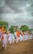 A group of people dressed in white uniforms are marching on a dirt ground holding Indian flags. The background features lush green trees under a cloudy sky.