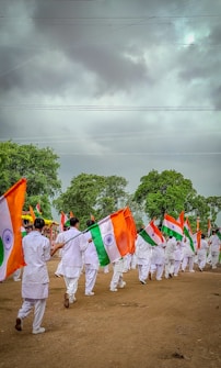 A group of people dressed in white uniforms are marching on a dirt ground holding Indian flags. The background features lush green trees under a cloudy sky.