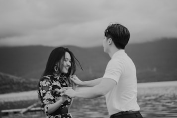 A black and white image captures two individuals smiling and holding hands in a playful manner by a body of water. The background features distant mountains under a cloudy sky, adding a serene and scenic element to the scene.