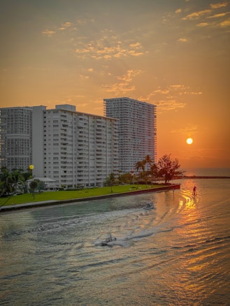 A picturesque sunset over a waterfront cityscape. Tall residential buildings stand beside a body of water, with a few boats moving across the calm surface. The sky is a warm orange, casting a golden reflection on the water's surface. Palm trees are visible along the green shoreline, adding a tropical feel to the urban environment.