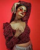 A person is posing confidently against a vibrant red background, wearing red-tinted sunglasses and a polka-dot top with puffed sleeves. The style suggests a bold and fashion-forward attitude.