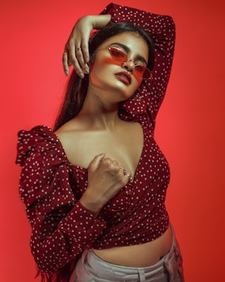 A person is posing confidently against a vibrant red background, wearing red-tinted sunglasses and a polka-dot top with puffed sleeves. The style suggests a bold and fashion-forward attitude.