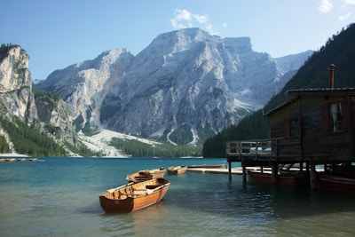 Wooden boats are gently floating near a rustic boathouse on a serene lake surrounded by towering mountains. The sunlight bathes the rugged landscape, casting shadows on the forest-covered slopes.