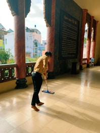 A person is mopping the floor of a spacious corridor with large columns and an inscription on the wall. There are trees and buildings visible through the open side of the corridor.