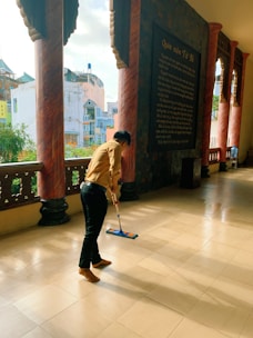 A person is mopping the floor of a spacious corridor with large columns and an inscription on the wall. There are trees and buildings visible through the open side of the corridor.