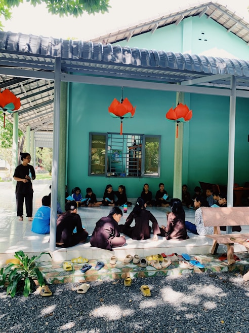 A group of individuals sit closely together on a tiled floor under a covered area, appearing to engage in a discussion or learning activity. The setting includes bright turquoise walls, a window with bars, and decorative orange lanterns hanging from the ceiling. Multiple pairs of shoes are neatly arranged on the ground at the entrance, and a bench is visible on the right side.