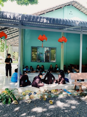 A group of individuals sit closely together on a tiled floor under a covered area, appearing to engage in a discussion or learning activity. The setting includes bright turquoise walls, a window with bars, and decorative orange lanterns hanging from the ceiling. Multiple pairs of shoes are neatly arranged on the ground at the entrance, and a bench is visible on the right side.