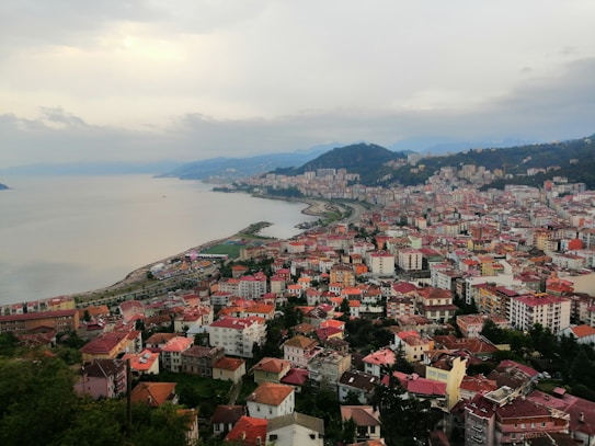 An urban coastal area is displayed with a densely built cityscape featuring numerous buildings with red rooftops and a mix of low-rise and high-rise structures. A large body of water borders the city on one side with a curved shoreline, and a range of green hills and mountains are visible in the background under a cloudy sky.