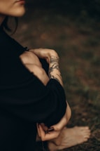 A person with a tattoo on their forearm is sitting with their knees drawn up. The focus is on the intricate tattoo design, which appears to be floral. The background is blurred, enhancing the intimate and serene atmosphere.