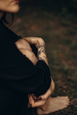 A person with a tattoo on their forearm is sitting with their knees drawn up. The focus is on the intricate tattoo design, which appears to be floral. The background is blurred, enhancing the intimate and serene atmosphere.
