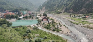 A scenic landscape featuring a river winding through a valley with lush green hills. In the foreground, multiple streams converge near a small settlement with colorful buildings. People are gathered near the water and scattered along the riverbanks, with a prominent, pointed-roof structure near the center.