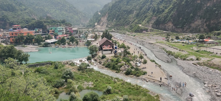 A scenic landscape featuring a river winding through a valley with lush green hills. In the foreground, multiple streams converge near a small settlement with colorful buildings. People are gathered near the water and scattered along the riverbanks, with a prominent, pointed-roof structure near the center.
