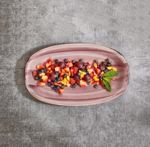 A colorful assortment of fruits including strawberries, blueberries, grapes, and diced mangoes presented on a rectangular, pastel-colored ceramic platter. A sprig of fresh mint adds a touch of green on the side. The platter is set against a textured gray background, creating a vivid contrast with the vibrant fruit colors.
