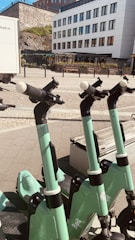 Several green electric scooters are lined up on a paved sidewalk in front of a modern white building with large windows. The background features a stone wall and some greenery. The sunlight casts shadows, creating a bright and clean urban setting.
