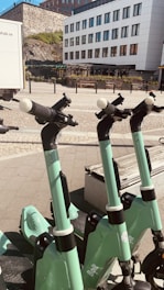 Several green electric scooters are lined up on a paved sidewalk in front of a modern white building with large windows. The background features a stone wall and some greenery. The sunlight casts shadows, creating a bright and clean urban setting.