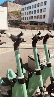 Several green electric scooters are lined up on a paved sidewalk in front of a modern white building with large windows. The background features a stone wall and some greenery. The sunlight casts shadows, creating a bright and clean urban setting.