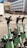 Several green electric scooters are lined up on a paved sidewalk in front of a modern white building with large windows. The background features a stone wall and some greenery. The sunlight casts shadows, creating a bright and clean urban setting.
