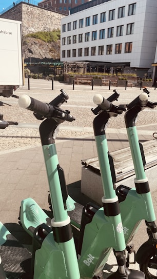 Several green electric scooters are lined up on a paved sidewalk in front of a modern white building with large windows. The background features a stone wall and some greenery. The sunlight casts shadows, creating a bright and clean urban setting.