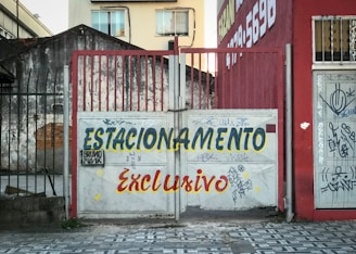A graffiti-covered gate in an urban setting displays the words 'ESTACIONAMENTO Exclusivo' in bold, colorful letters. The background features a weathered building facade with visible graffiti and nearby a well-worn sidewalk.