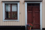 A wooden exterior wall painted in a light beige with vertical paneling features a large window framed in white and brown to the left. Next to the window is a solid brown door with rectangular panels, also framed in white. In front of the door is a terracotta pot containing colorful flowers, notably red blooms and green foliage.