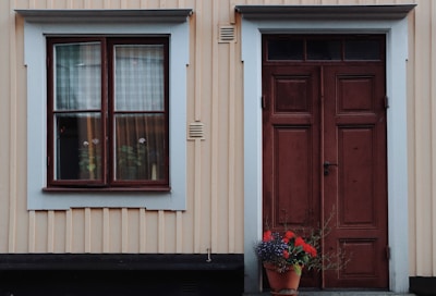 A wooden exterior wall painted in a light beige with vertical paneling features a large window framed in white and brown to the left. Next to the window is a solid brown door with rectangular panels, also framed in white. In front of the door is a terracotta pot containing colorful flowers, notably red blooms and green foliage.