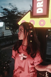 A young person with long dark hair adorned with colorful flowers is standing near a large illuminated yellow arrow sign that reads 'DR'. The person is wearing a red t-shirt with white and black text on it. The background features trees and an urban setting, with a dim evening light providing a moody atmosphere.