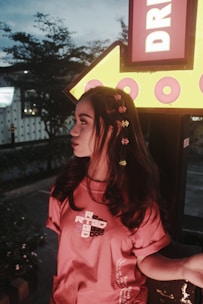 A young person with long dark hair adorned with colorful flowers is standing near a large illuminated yellow arrow sign that reads 'DR'. The person is wearing a red t-shirt with white and black text on it. The background features trees and an urban setting, with a dim evening light providing a moody atmosphere.