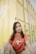 A young woman sits in front of a yellow industrial container. She wears a red-orange t-shirt with a graphic design and khaki pants. Her long dark hair is adorned with small flower clips. The expression on her face is calm and composed.