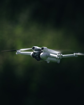 A white drone with four propellers is captured mid-flight against a blurred, dark green background. The drone is centrally composed, with its camera and sensors clearly visible, giving an impression of precision and advanced technology.