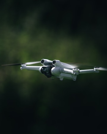 A white drone with four propellers is captured mid-flight against a blurred, dark green background. The drone is centrally composed, with its camera and sensors clearly visible, giving an impression of precision and advanced technology.