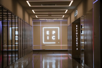 A corridor with reflective floors leads to a wall illuminated with a large neon sign displaying the letters 'E P I C'. The walls are lined with mirrored surfaces, and there is a vertical sign displaying 'REVOLUCNI 3'. The ceiling features modern, recessed lighting.