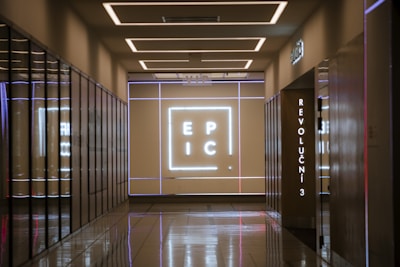 A corridor with reflective floors leads to a wall illuminated with a large neon sign displaying the letters 'E P I C'. The walls are lined with mirrored surfaces, and there is a vertical sign displaying 'REVOLUCNI 3'. The ceiling features modern, recessed lighting.