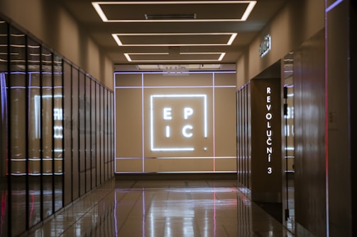 A corridor with reflective floors leads to a wall illuminated with a large neon sign displaying the letters 'E P I C'. The walls are lined with mirrored surfaces, and there is a vertical sign displaying 'REVOLUCNI 3'. The ceiling features modern, recessed lighting.